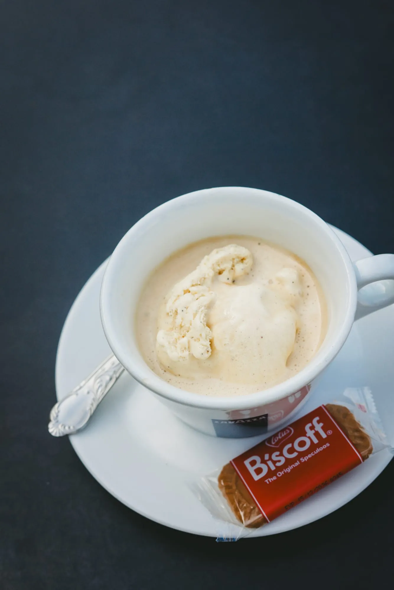 Vue de dessus : une tasse de café avec une boule de glace, une cuillère et un biscuit Biscoff, sur une table sombre.