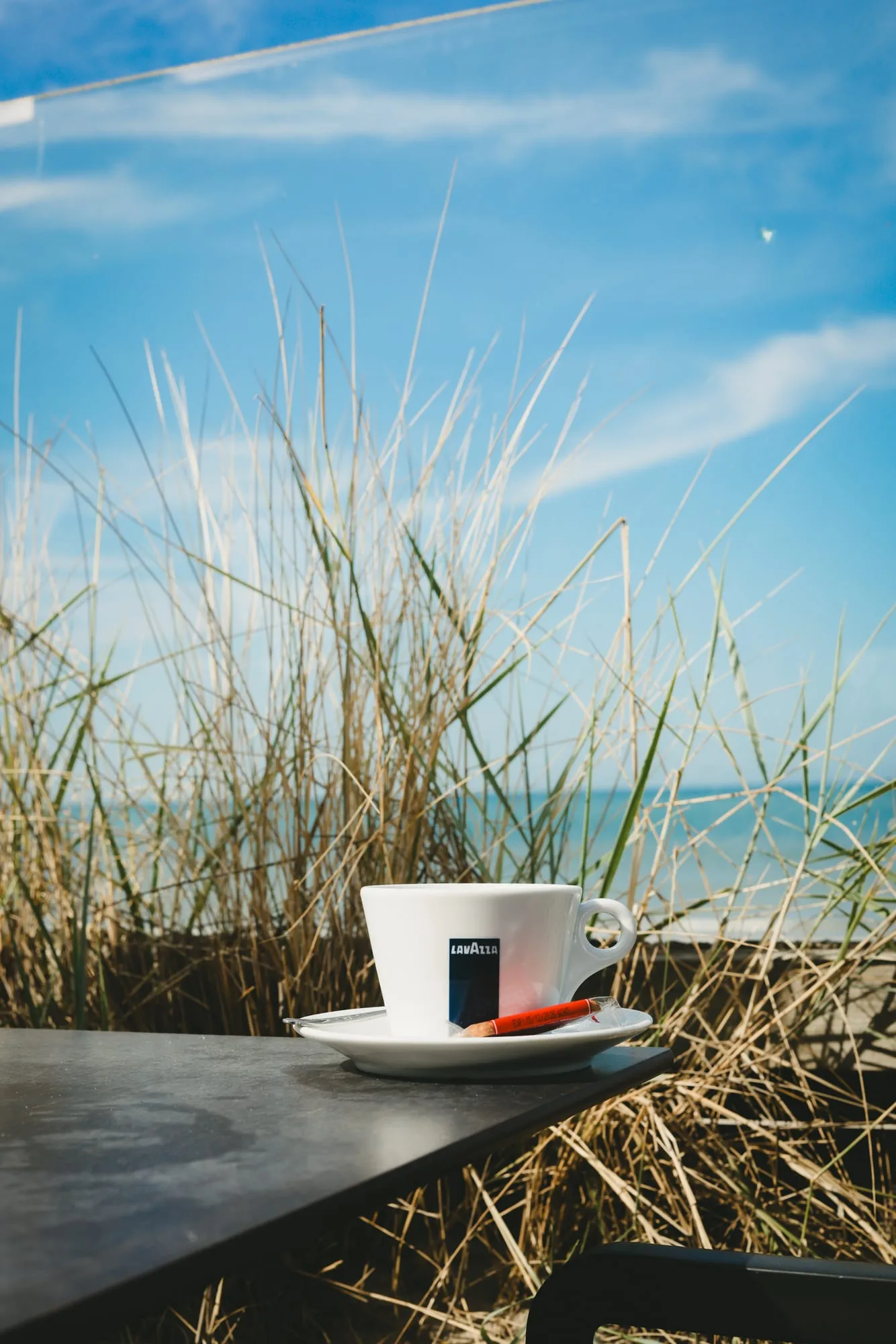 Une tasse de café posée sur une table, au milieu des oyats, la mer derrière.