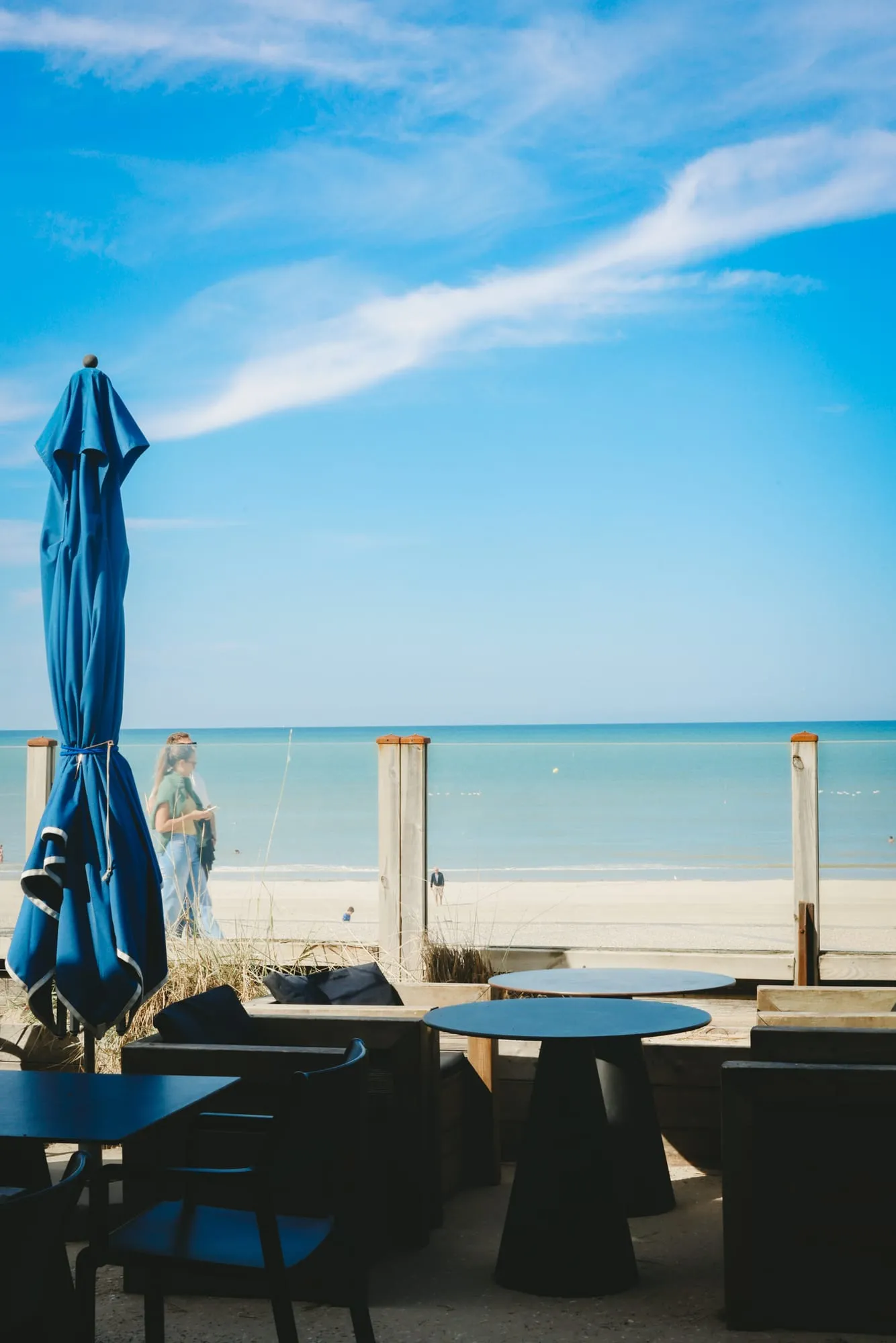 La terrasse face à la mer : un parasol bleu replié, des tables rondes, une promeneuse sur la digue.
