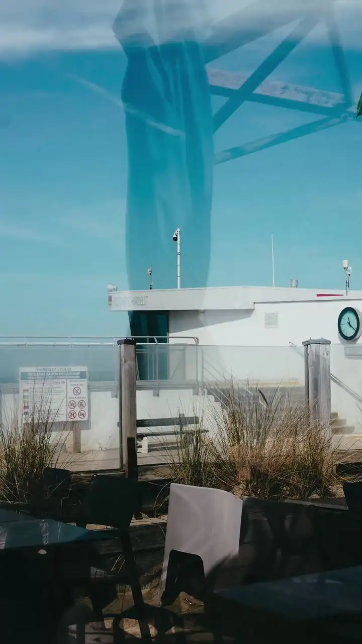 Depuis la salle, à travers la vitre : le reflet d’un parasol bleu, le poste de secours et son drapeau vert.